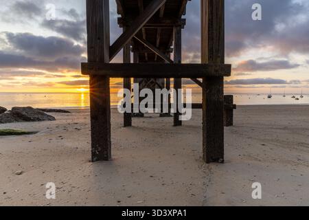 Un'alba sul mare si illumina attraverso i sostegni di legno intemprati di un Plage des Dames, molo sulla spiaggia, che getta lunghe ombre su sabbia liscia. Le barche navigano calme W Foto Stock