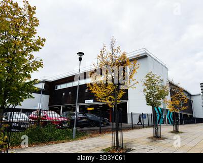 Manchester, Regno Unito - 30 settembre 2025: Edificio moderno con parcheggio, alberi autunnali e pedoni a Manchester, Regno Unito. Foto Stock
