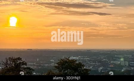 Il vivace tramonto proietta tonalità calde su un vasto skyline cittadino, con nuvole che riflettono la luce dorata e sagome di alberi che incorniciano la scena, creando Foto Stock