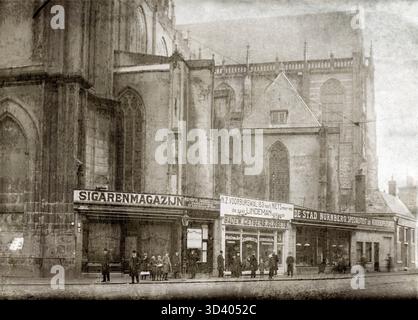 Il Nieuwe Kerk sulla diga di Amsterdam, con negozi alla base che vendono sigari e sanitari e pedoni a piedi. L'immagine mostra una tipica scena urbana di Amsterdam nel 1909. Paesi Bassi, 1909 Foto Stock