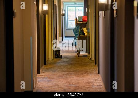 Un lungo corridoio dell'hotel mostra le porte delle camere e un grande carrello per le pulizie impilato con asciugamani, carta igienica e sacchetti della spazzatura, la sala tranquilla sembra ordinata Foto Stock