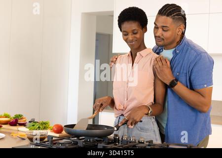 Cuocere diverse coppie mescolando cibo su fornelli a gas in una cucina luminosa, con spatola e verdure Foto Stock
