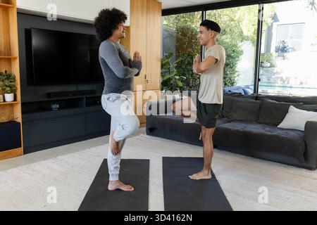 Praticare la posa di alberi diversi amici maschi che si bilanciano su tappetini da yoga a casa, con TV e piante Foto Stock