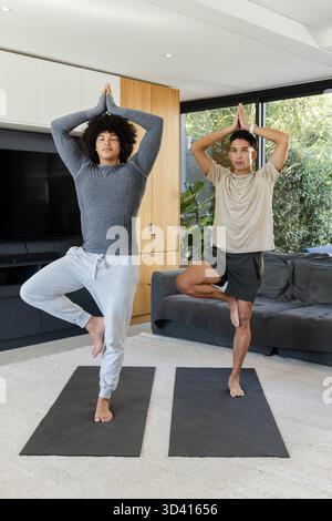 Praticando diversi amici maschi che si esibiscono in posa sull'albero nel moderno soggiorno, con stuoie per yoga e TV Foto Stock