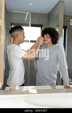 Applicazione di patch in idrogel rosa, amici diversi in piedi al lavandino del bagno con specchio e asciugamano Foto Stock