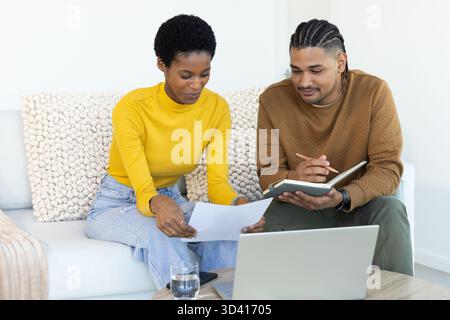 Coppia diversificata che rivede documenti e laptop sul divano in soggiorno, con smartphone Foto Stock