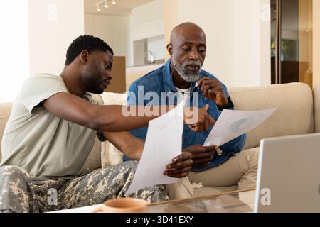 Famiglia afroamericana seduta che esamina le carte stampate sul divano beige, con laptop e tazza da caffè Foto Stock