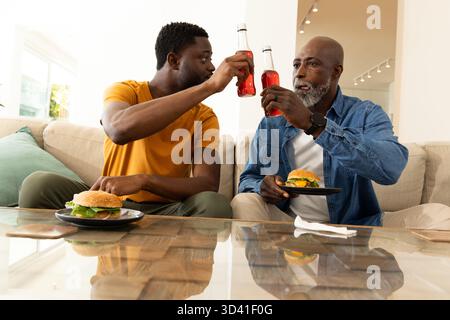 Tostare padre e figlio afroamericani seduti sul divano a casa, tenendo hamburger e bottiglie rosse Foto Stock