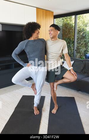 Bilanciare diversi amici maschi che praticano la posa degli alberi in salotto, con tappetini per yoga e smartwatch Foto Stock