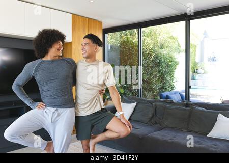 Bilanciare diversi amici maschi che praticano yoga in coppia nel soggiorno, con TV a schermo piatto Foto Stock