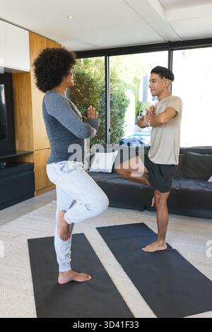 Bilanciare diversi amici maschi che praticano posa sugli alberi su tappetini da yoga nel soggiorno, con porte in vetro Foto Stock