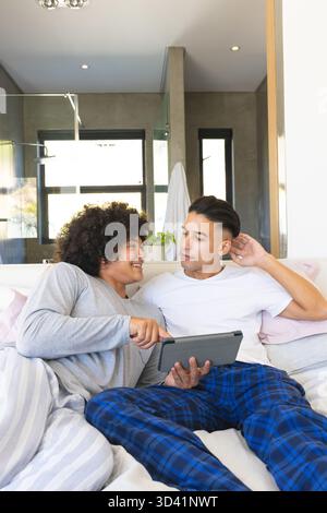 Sorridere diversi amici maschi che condividono un tablet sul letto in una casa moderna, con cuscini decorativi Foto Stock