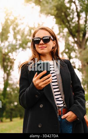 Ritrattista che indossa occhiali da sole e cappotto nero con smartphone nel parco autunnale. Foto Stock