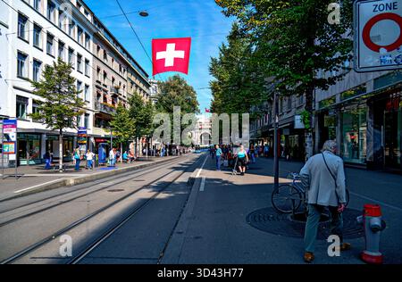 La bandiera svizzera è appesa sopra la famosa via dello shopping "Bahnhofstrasse" con vista sulla stazione ferroviaria principale di Zurigo, Svizzera; Foto Stock
