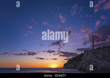 Statua obelisco e bandiera montenegrina che fluttua nel vento nella fortezza sulla cima della scogliera a Petrovac costa al tramonto, Montenegro Foto Stock