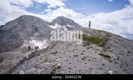 Uomo sulla collina che guarda su una capanna alpina con montagne sullo sfondo, Parco Nazionale del Triglav, Slovenia Foto Stock