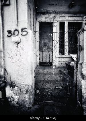Immagine in bianco e nero di un vecchio ingresso con cancello metallico, scale e muro marcato di un edificio urbano deteriorato Foto Stock