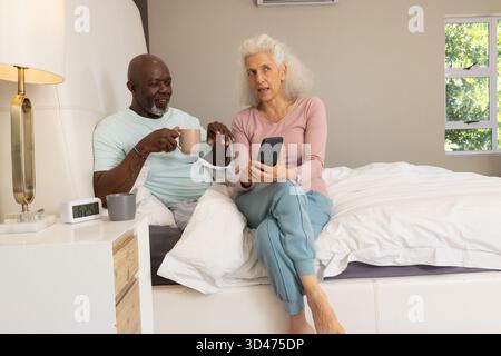Rilassanti coppie anziane che controllano lo smartphone sul letto, con tazze da caffè, cuscini e lampada Foto Stock