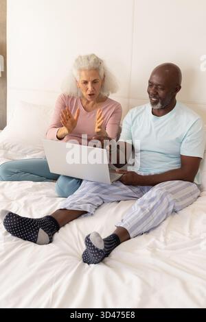 Condividere foto di una coppia senior utilizzando un computer portatile color argento sul letto in camera da letto, con gesti delle mani Foto Stock