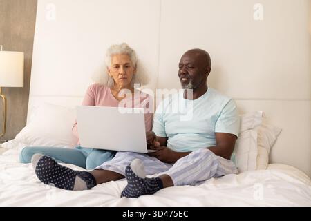 Diverse coppie anziane sedute sul letto usando un computer portatile color argento in una camera da letto luminosa, con piantana color oro Foto Stock
