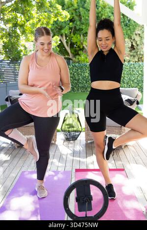 Diverse amiche che praticano yoga bilanciato sul patio sotto il pergolato, con supporto per illuminatore anulare Foto Stock