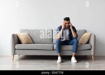 Il giovane arabo seduto sul divano soffre di forte mal di testa e stress mentre tocca i suoi templi Foto Stock