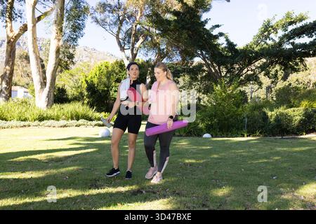 Cammina con diverse amiche che chiacchierano sul prato erboso del parco, tenendo in mano tappetini e asciugamani per lo yoga Foto Stock