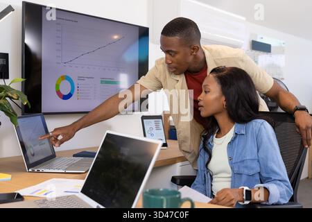 Collaborazione con colleghi afroamericani che analizzano grafici su laptop in un ufficio moderno, con MUG Foto Stock