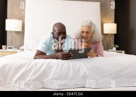 Sdraiato da coppie anziane diverse che si appoggiano sui gomiti in una camera da letto moderna, sfogliando i contenuti su tablet Foto Stock