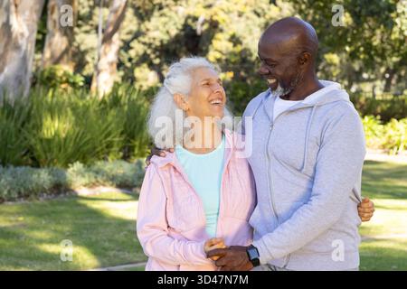 Coppia sorridente che tiene le mani in un parco illuminato dal sole vicino al sentiero, mostrando uno smartwatch Foto Stock