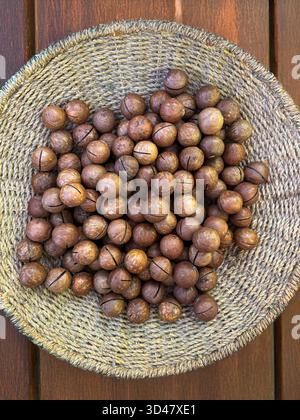 Nocciole di macadamia intere in un cestino intrecciato su sfondo di legno Foto Stock