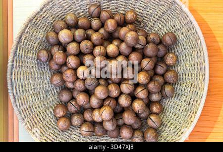 Nocciole di macadamia intere in un cestino intrecciato su sfondo di legno Foto Stock
