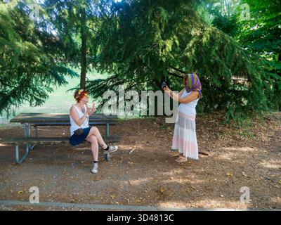 Due giovani donne eleganti che si godono una giornata di sole all'aperto scattando foto l'una dell'altra su una panchina del parco circondata da lussureggianti alberi verdi e luce naturale Foto Stock