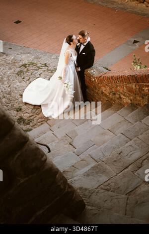 La sposa e lo sposo condividono un bacio tenero su gradini di pietra, circondati da un ambiente rustico. La sposa regge un bouquet di fiori bianchi, il suo elegante abito flowi Foto Stock