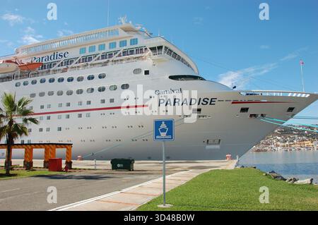 Grande nave da crociera bianca ormeggiata in un porto soleggiato, mostrando la sua prua e i ponti multipli, con un cartello nautico, prato verde e città costiera nel backgrou Foto Stock