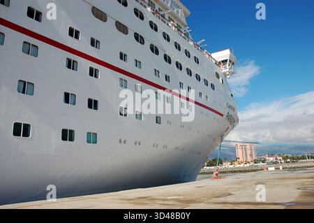 Grande nave da crociera bianca ormeggiata in un porto soleggiato, mostrando la sua prua e i ponti multipli, con un cartello nautico, prato verde e città costiera nel backgrou Foto Stock