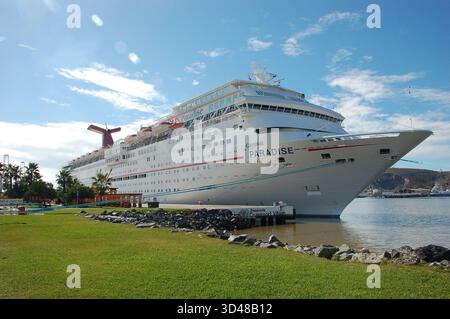 Grande nave da crociera bianca ormeggiata in un porto soleggiato, mostrando la sua prua e i ponti multipli, con un cartello nautico, prato verde e città costiera nel backgrou Foto Stock