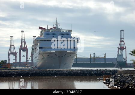 Grande nave da crociera bianca ormeggiata in un porto soleggiato, mostrando la sua prua e i ponti multipli, con un cartello nautico, prato verde e città costiera nel backgrou Foto Stock
