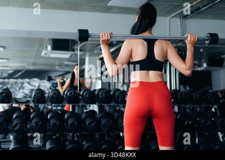 Vista posteriore di una bella donna sportiva che fa squadra con barbell al club fitness. Giovane donna dai capelli scuri con top nero, pantaloncini rossi e bianco Foto Stock