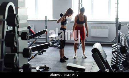 Vista laterale di una donna magra felice in un allenamento sportivo con barbell, mentre l'altra donna in piedi prima e aiutandola. Giovane donna che si esercita regolarmente Foto Stock