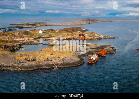 Un relitto intempestivo si trova sotto la superficie, accanto a una suggestiva boathouse rossa su una piccola isola, incorniciata dalla spettacolare costa della Norways Atlantic Ocean Road. Foto Stock