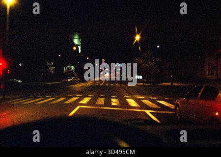 Pilota notturno POV sopra un passaggio pedonale in un incrocio, in stile pellicola analogica sgranata che cattura le luci urbane e la prospettiva. Foto Stock