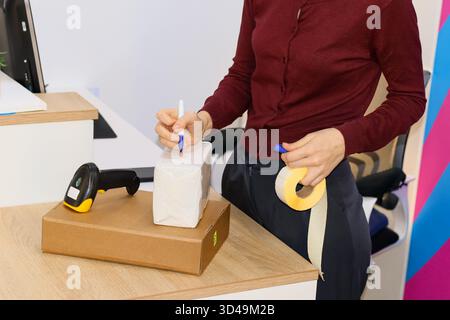 Etichettare il pacco dei lavoratori al banco di ritiro della consegna. Il dipendente in maglione marrone scrive su un articolo avvolto in bianco sopra una scatola marrone con scanner Foto Stock