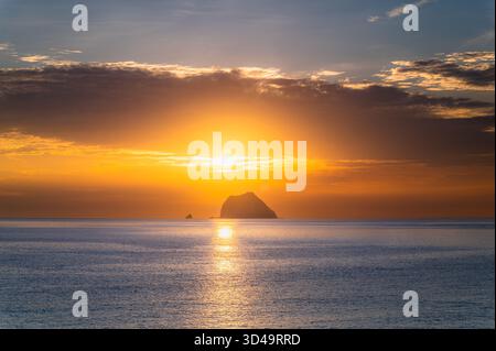 Il sole sorge attraverso le spettacolari nuvole dietro l'isolotto di Keelung, visto dalla costa di Waimushan, Taiwan, che proietta luce dorata e arancione sull'oceano calmo. Foto Stock