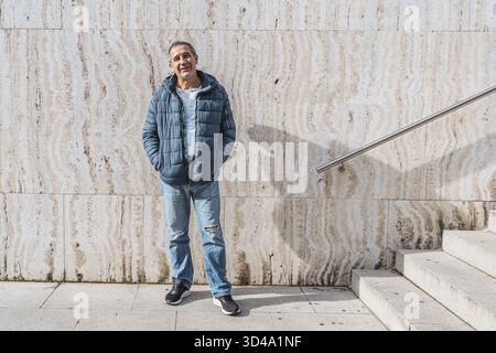 Ritratto uomo di mezza età allegro e positivo, in giacca blu caldo, jeans strappati alle ginocchia, vicino al muro di marmo, su gradini con corrimano Foto Stock
