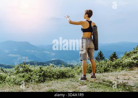 Una giovane donna, atletica e attraente, guarda le montagne dal retro mentre si trova su un'alta roccia Foto Stock