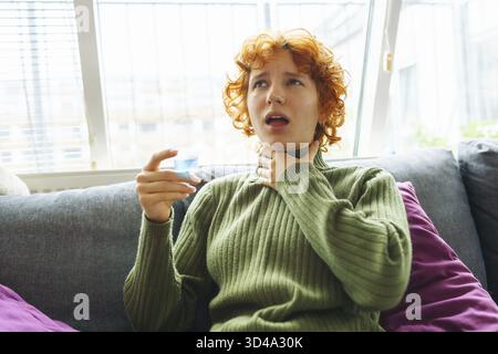Ritratto di giovane donna dai capelli rossi in maglione verde lana caldo, coperto da una coperta calda, prende medicine, infezione da freddo gola Foto Stock