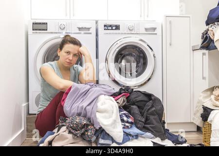 Turbare la donna nella sala lavanderia seduta ona piano con vestiti sporchi, stanchi e frustrati Foto Stock