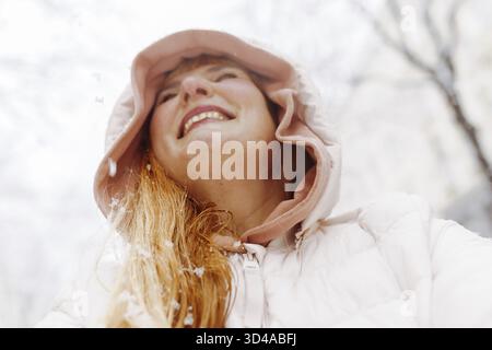 Ritratti una donna di mezza età positiva con lunghi capelli biondi, indossa una giacca calda con cappuccio e scatta gioiosamente selfie sulla neve per strada europea Foto Stock