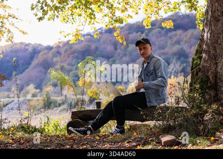L'uomo sorseggia un caffè contento con un notebook nei boschi autunnali. L'uomo sorridente tiene la tazza di caffè alle labbra, il laptop in grembo mentre è seduto sul tronco all'aperto Foto Stock